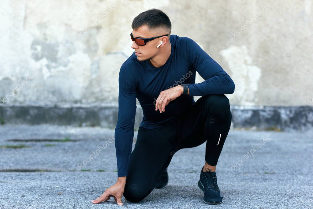 Horizontal image of a sporty male resting outdoors after the workout. Fitness man in blue sports clothing and eyewear exercising with earphones outside against the urban concrete wall.
