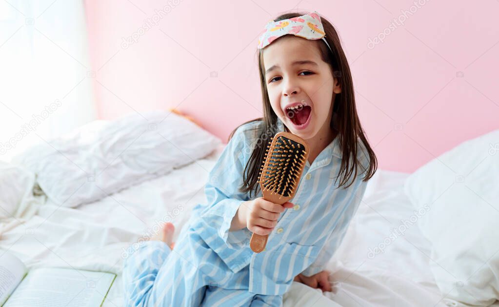 Portrait of a joyful little girl in pajama holding a hair brush like microphone singing imitates herself a real singer in the morning at home.