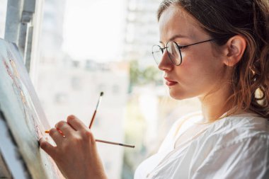Resim stüdyosundaki tuvale resim çizen güzel bir kadın ressamın yakından yan görüntüsü pencerenin yanında oturuyor. Yağlı gözlüklü bir kadın ressam atölyede hayal gücü arıyor.