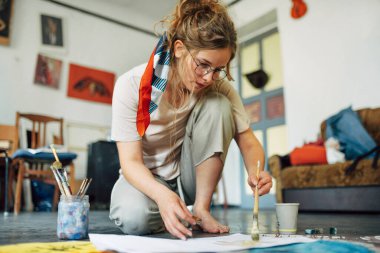 Sanat stüdyosunda yerde oturup kağıt üzerinde resim yapan güzel bir kadın sanatçının yatay görüntüsü. Atölyede suluboya gözlüklü bir kadın ressam..