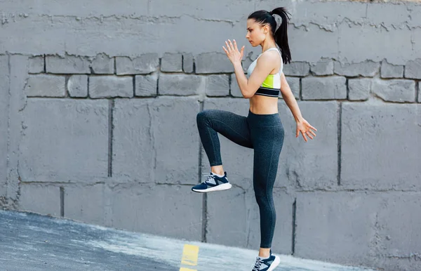 运动员年轻女子在混凝土灰墙上跳跃运动的横向侧视图 早上在外面灰色的墙上锻炼时 白种人的女性穿着时髦的运动服 — 图库照片