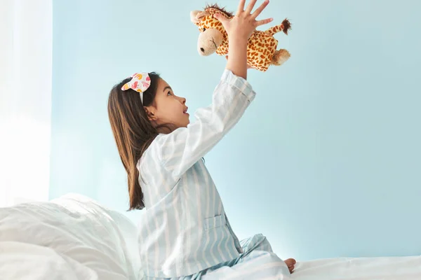 Cute little girl wearing pajama playing with toy giraffe in the bed, isolated on light blue background. Child playing in the morning in her bedroom.