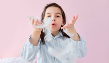 Pembe arka planda, yatakta oynarken pijamalı, beyaz tüyler üfleyen esmer bir kızın yakın çekimi. Sabahları oyun oynayan sevimli bir çocuk..