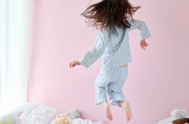 Mavi pijamalı esmer bir kızın sabahları güneşli, modern bir odada dans edişinin arka görüntüsü. Mutlu çocuk sabah evde doğum gününde yatakta zıplıyor ve dans ediyor..