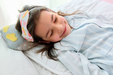 Çizgili mavi beyaz pijama giyen, uyku maskesi takan, gözleri kapalı gülümseyen ve yatakta dinlenen sevimli küçük bir kızın portresi. Çocuğun sabah neşeli bir ifadesi var..