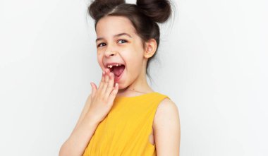 Sarı elbiseli neşeli anaokulu kızı, gri stüdyo arka planında izole edilmiş ilk azı dişini büyütürken kendini neşeli hissediyordu. İlk bebek dişini kaybeden sevimli bir çocuk. Çocuk süt dişini kaybetti..