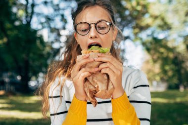 Güzel bir kız öğrencinin portresi açık havada sandviçi ısırıyor. Şeffaf gözlüklü muhteşem sarışın bir kadın parkta fast food yemeye ara veriyor..