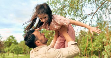 Baba parkta kızını kollarıyla kaldırıyor. Baba ve çocuk güneşli bir günde parkta oynuyorlar..