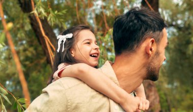 Neşeli kız babasının sırtında gezerken kendini mutlu hissediyor. Baba, güneşli bir günde çocuğunu sırtında taşıyarak parkta yürüyor..