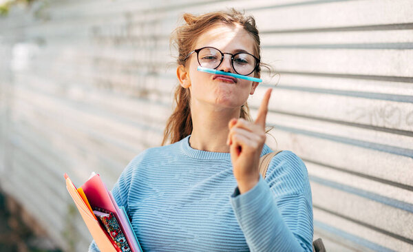 A funny female student in transparent eyeglasses carrying lots of books after a day in the college and making grimace with a pencil posing outdoors.