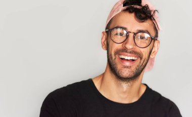 Gülümseyen, doğrudan kameraya bakan, reklam için poz veren, siyah tişört, pembe şapka ve şeffaf gözlük takan, gri stüdyo arka planında izole edilmiş genç bir adamın yakın plan portresi..