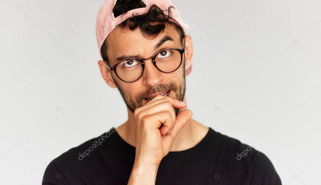 Closeup portrait of a doubtful young man doesn't know what to do, dressed in stylish black t-shirt, transparent eyeglasses, and pink cap. Male has confused expression.