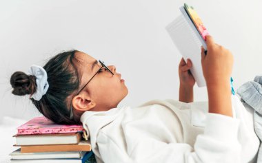 Üniformalı, şeffaf gözlüklü, kitap yığınının üzerinde uzanmış kitap okuyan küçük bir kızın yan görüntüsü. Liseli kız gri stüdyo arkaplanına karşı bir sürü kitap okuyor..