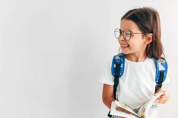 透明な眼鏡の中の少女の水平方向のイメージで バックパックのコピースペースで片側を見て笑っている バックパックと本を持つ学校の子供は学校に戻る喜びの表現を持っています — ストック写真