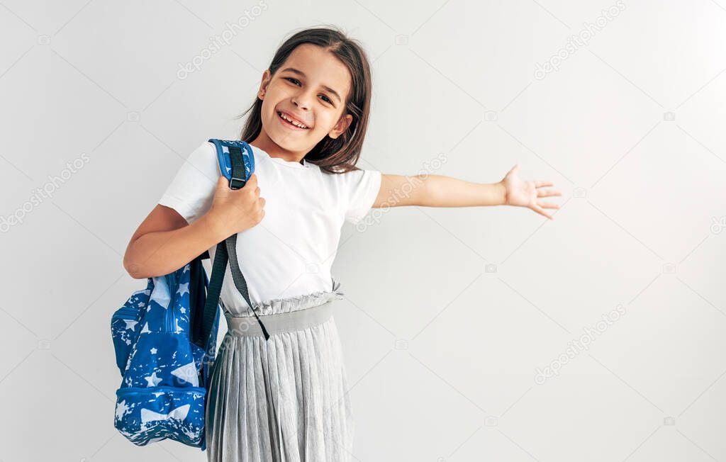 Happy schoolgirl with blue backpack smiling and looking at camera. Pretty school kid with backpack has joyful expression with arm wide open. Studio portrait of little girl posing over grey background.