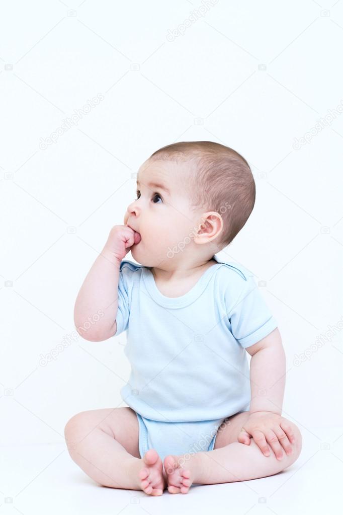 Look at one side and sitting on white background beautiful baby with hand in mouth