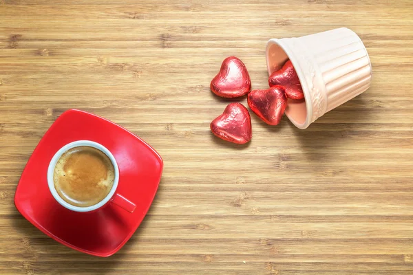 Dulces en forma de corazón envueltos en una lámina de color rojo brillante que yace en un jarrón de cerámica sobre una textura de madera con taza de café rojo cerca de ella. Fondo para temas románticos . — Foto de Stock