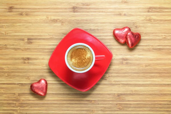 Szív alakú édesség fóliába csomagolva egy fényes vörös feküdt egy fa szerkezetű piros csésze kávé közelében. Háttérben a romantikus témák. — Stock Fotó