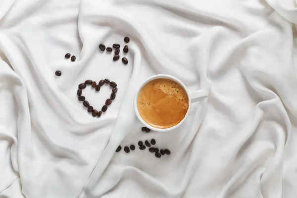 Taza blanca de café sobre una tela de seda blanca. Taza de café expreso. Granos de café esparcidos en forma de corazón sobre una tela de seda blanca. Vista superior. Desayuno romántico para San Valentín . — Foto de Stock