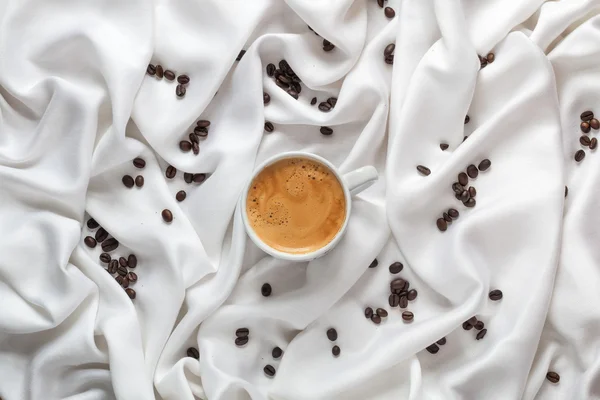 Taza blanca de café sobre una tela de seda blanca. Taza de café expreso con espuma en forma de cara sonriente. Granos de café esparcidos en una tela de seda blanca. Vista superior . — Foto de Stock