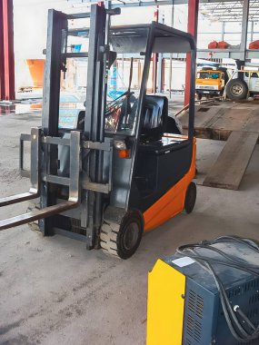 An orange and black forklift stands in a warehouse, ready for work. Other vehicles are visible in the background, along with a charging station.