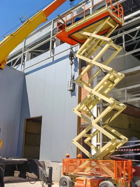A construction scene features a yellow scissor lift and crane working on a building under a bright blue sky. Workers are present.