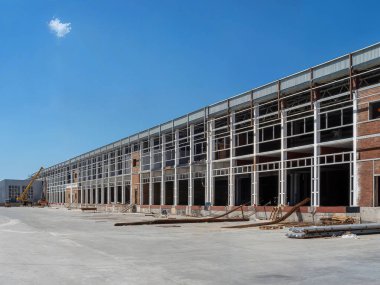 Uzun bir sanayi binası inşaat halinde, çelik çerçeveler ve tuğla duvarlar korunmasız durumda. Gökyüzü açık mavi ve bulutlu..