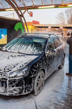 A black station wagon is covered in soap suds at a car wash, undergoing a thorough cleaning process by a person.