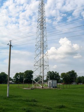 A tall metal communication tower stands prominently in a green field, with power lines stretching across a partly cloudy sky.  Trees and a small building surround it.