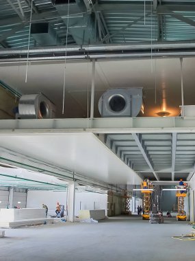 İşçiler tavan panelleri kurarken ve büyük bir deponun havalandırma sistemi üzerinde çalışırken görülüyor.