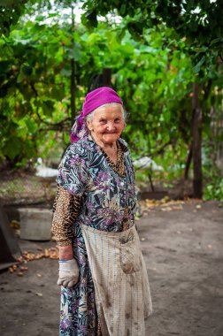 Yaşlı bir kadın başörtüsü ve önlükle dışarıda dikilirken sıcak bir şekilde gülümsüyor ve doğrudan kameraya bakıyor.