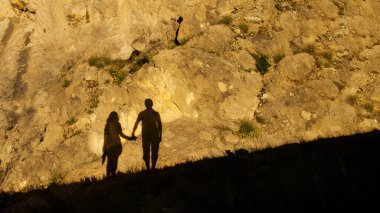 Birlikte yürüyen bir çiftin silüetleri, dokulu bir kaya yüzüne karşı romantik bir sahne yaratıyor.