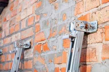 Metalik montaj donanımının yakın görüntüsü sert tuğla bir duvara sabitlenmiş, devam eden inşaat veya onarım çalışmalarını gösterir