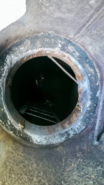 Endüstriyel bir yapının derin, karanlık, dairesel açılışına yukarıdan bakıldığında bilinmeyen bir yere inen metal bir merdiven görülüyor.