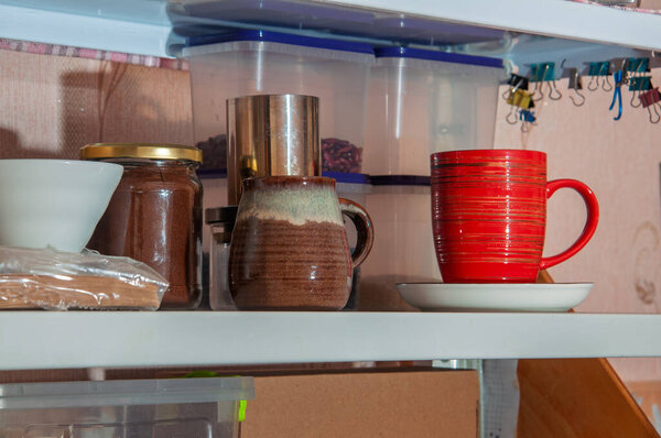 A close-up view of a kitchen shelf displaying various items including a jar of coffee grounds, two mugs, and clear plastic food storage containers