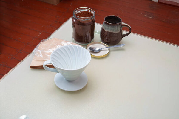 A collection of items for brewing coffee, including a pour-over dripper, coffee grounds in a jar, filters, and a mug, are arranged on a light-colored surface