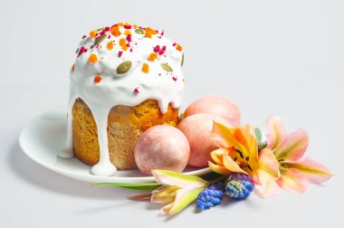 Pastel renkli yumurtalar ve beyaz bir yüzey üzerinde sunulan narin çiçeklerle çevrili, krema ve malzemelerle süslenmiş neşeli bir Paskalya kulişi.