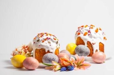 Beyaz krema ve şekerli meyvelerle süslenmiş iki güzel Paskalya pastası renkli boyanmış yumurtaların ve narin bahar çiçeklerinin yanında oturur.
