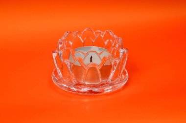 Nilüfer çiçeği şeklinde berrak bir cam adak tutucu, parlak turuncu bir arka fona karşı uyumlu bir fincan tabağının üzerinde oturmakta, bir uzayı aydınlatmaya hazır.