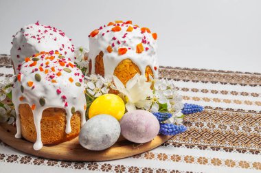 Yakın çekimde, beyaz krema, kurutulmuş meyve ve tohumlarla süslenmiş üç güzel dekore edilmiş Paskalya pastasının yanında renkli boyanmış yumurtalar ve narin bahar çiçekleri sergileniyor.