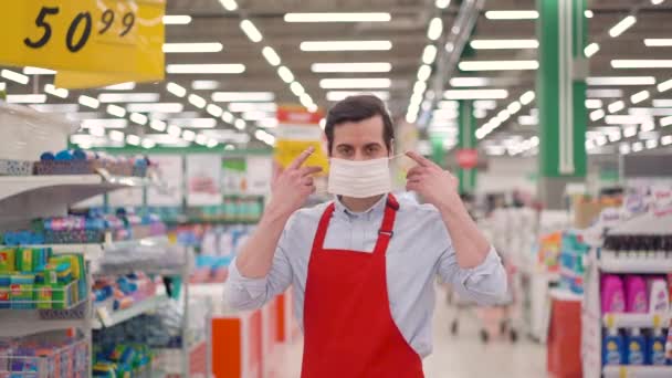 Голова знімає молодого кавказького продавця в червоному фартусі одягає білу захисну медичну маску, що стоїть в супермаркеті з фоном полиць, дивлячись на камеру. Припинити концепцію пандемії вірусу корона — стокове відео