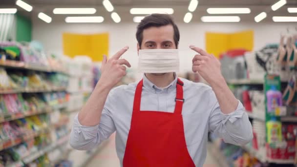 Head shot young caucasian seller man in red apron puts on white protective medical face mask standing posing on grocery store background looking at camera. Stop corona virus pandemic outbreak concept — Stock Video