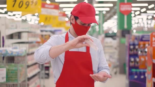 Hezký kavkazský mladý prodejce nosí červenou masku a čistící ruce s dezinfekčním gelem v supermarketu. Zaměstnanec používající dezinfekci alkoholu před prací na prevenci koronavirového konceptu. — Stock video