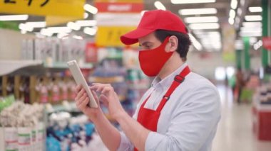 Beyaz erkek mağaza çalışanı, kırmızı maskeli ve üniformalı süpermarkette duruyor ve tablet bilgisayarda yazıyor. Salgın covid-19, koronavirüs salgını sırasında teslimat servisinde çalışan satıcı.