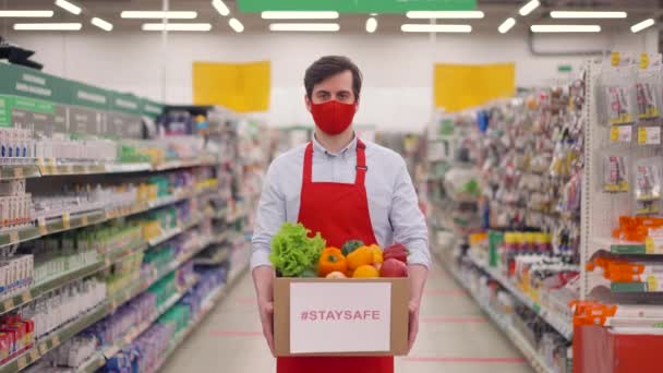 Bonito entregador de máscara vermelha em pé no supermercado com legumes em caixa de papelão. Vendedor trabalhando no serviço de parto durante epidemia covid-19, pandemia de coronavírus, compras online de alimentos — Vídeo de Stock