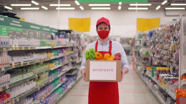 フェイスマスクの配達人と段ボール箱の野菜とスーパーマーケットで一様立って、流行時に配達サービスで働く宅配便covid-19 、コロナウイルスパンデミック、オンライン食品ショッピング — ストック動画