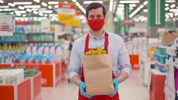 紙のパッケージに野菜とスーパーマーケットに立って赤い顔のマスクの男性の宅配便。流行のcovid-19の間の配達サービスで働く配達業者、コロナウイルスのパンデミック、オンライン食品ショッピング — ストック動画