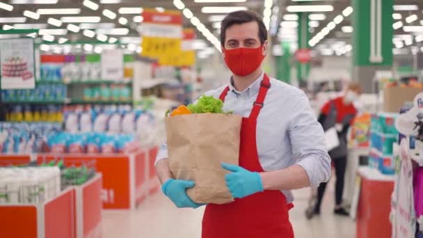 Distribuidor de tienda masculina en máscara facial roja de pie en el supermercado con comida en paquete de papel. Mensajero trabajando en el servicio de entrega durante epidemia covid-19, pandemia de coronavirus, compras de alimentos en línea — Vídeos de Stock