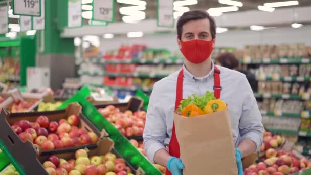 Чоловічий кур'єр в червоній масці стоїть в продуктовому магазині з овочами в паперовій упаковці. "Делівері" працює в службі доставки під час епідемії ковідо-19, пандемії коронавірусу, інтернет-магазинів продовольства — стокове відео