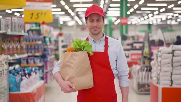 Sorrindo entregador masculino empregado vestindo uniforme vermelho segurando pacote de papel artesanal com legumes frescos em pé no supermercado. Rápido expresso e seguro entrega de alimentos compras on-line, conceito de comércio eletrônico — Vídeo de Stock
